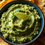 A close-up photograph of a dark blue bowl filled with creamy, textured guacamole, garnished with a slice of fresh lime and a sprig of mint. The bowl is surrounded by golden, crispy tortilla chips on a dark surface.