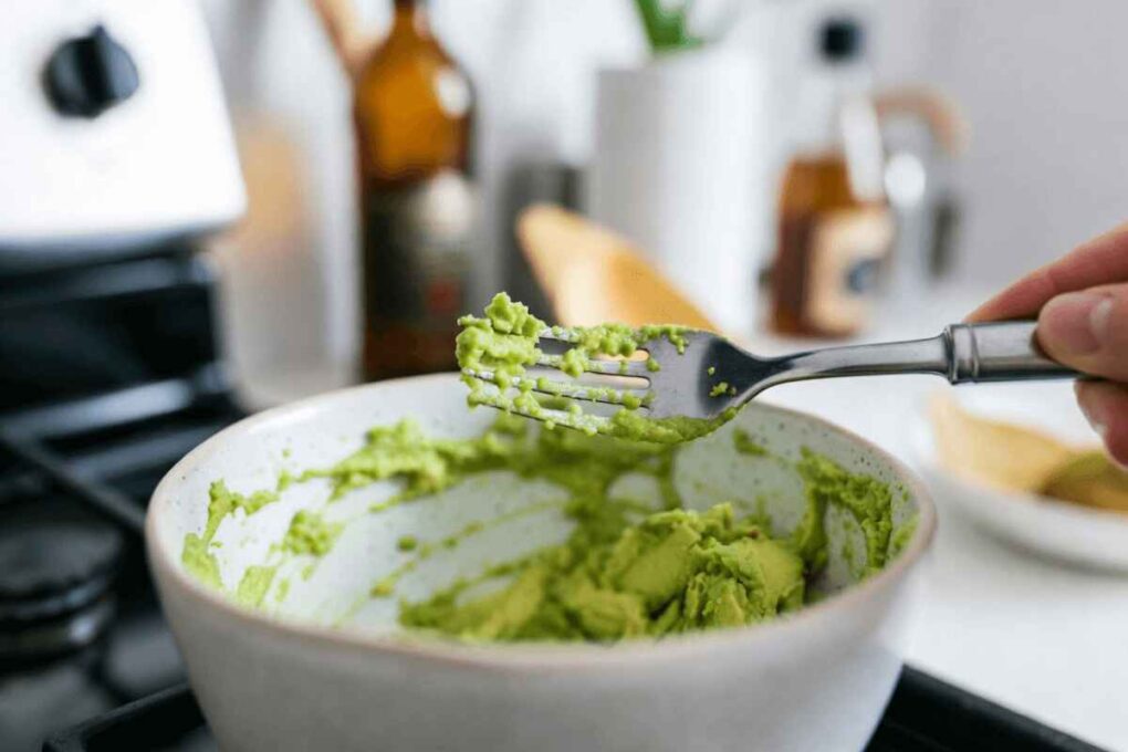 Close-up of a hand mashing ripe avocado with a fork in a white bowl while preparing a guacamole recipe in a kitchen.