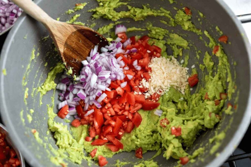 Close-up of a bowl filled with mashed avocado topped with chopped red onions, diced tomatoes, and minced garlic, with a wooden spoon ready to mix for a guacamole recipe.