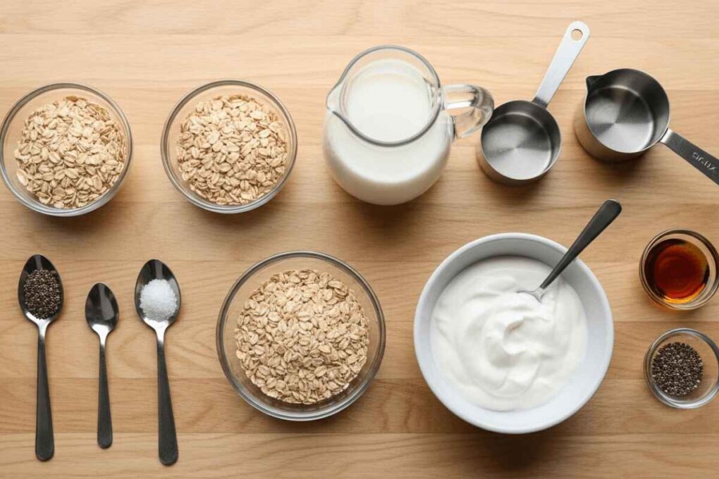 Overnight oats recipe ingredients laid out on a wooden surface including rolled oats, milk, Greek yogurt, chia seeds, salt, and maple syrup