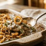 A spoonful of creamy green bean casserole topped with golden crispy fried onions being lifted from a rustic ceramic baking dish