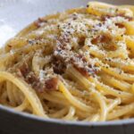 Close-up photo of creamy spaghetti carbonara served in a rustic ceramic bowl, topped with grated Pecorino Romano, cracked black pepper, and crispy halal beef bacon on a wooden table.
