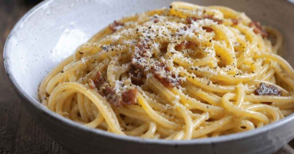 Close-up photo of creamy spaghetti carbonara served in a rustic ceramic bowl, topped with grated Pecorino Romano, cracked black pepper, and crispy halal beef bacon on a wooden table.