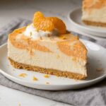 Slice of No-Bake Orange Creamsicle Cheesecake with creamy orange and vanilla layers, graham cracker crust, whipped cream, mandarin orange segments, and fresh orange zest.