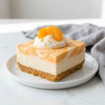 Square slice of orange creamsicle cheesecake with a graham cracker crust, topped with whipped cream, mandarin orange segments, and orange zest on a white plate.
