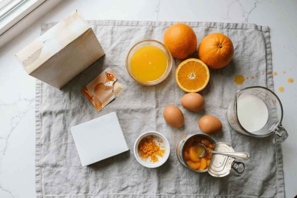 Orange Creamsicle Poke Cake – Moist, Easy, and Worth the Wait 6 Orange creamsicle poke cake ingredients flat lay: cake mix, gelatin packet, eggs, orange juice, fresh oranges and mandarin segments