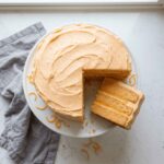 An overhead view of a frosted Orange dreamsicle cake on a white stand, with one slice pulled out to reveal three soft sponge layers and bright orange curd filling.